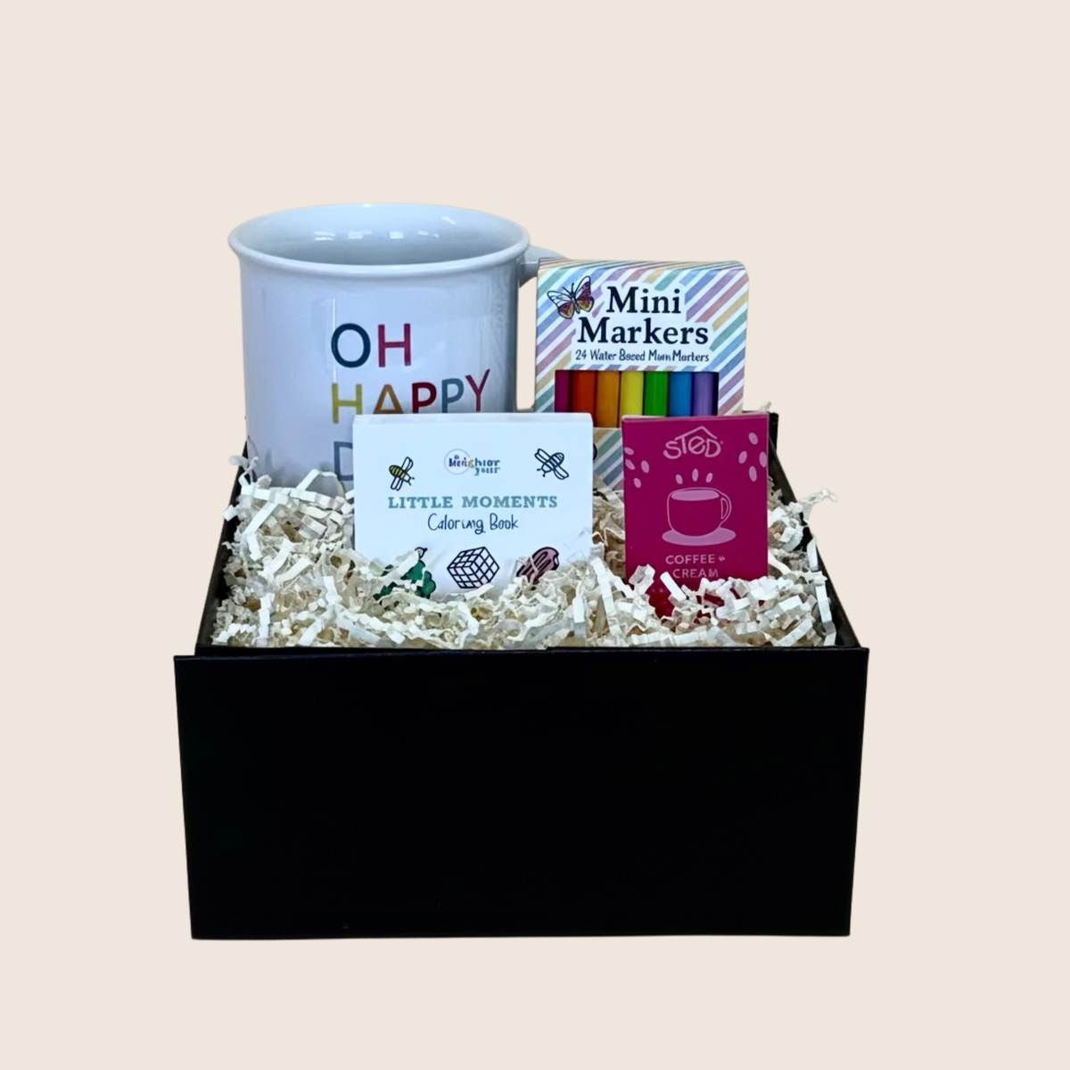 A coffee-themed gift box set in a black container filled with shredded paper. The set includes a white ceramic mug with 'OH HAPPY' printed on it, a pack of mini markers, a 'Little Moments Color-My Book', and a pink 'Coffee Lover' card.