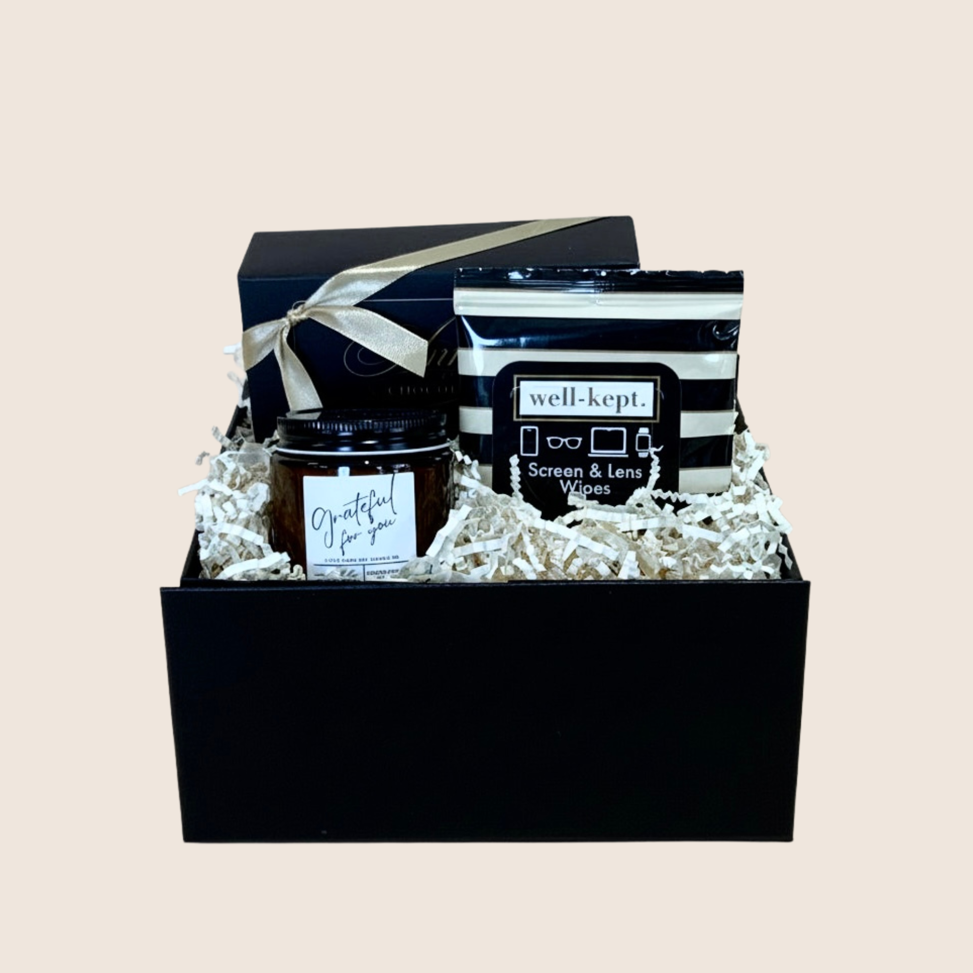 A curated gift box set containing a black jar candle labeled 'grateful for you', a black-and-white striped bag of 'well-kept. Screen & Lens Wipes', and a black gift box tied with a beige ribbon, all arranged in white crinkle paper on a light beige background.