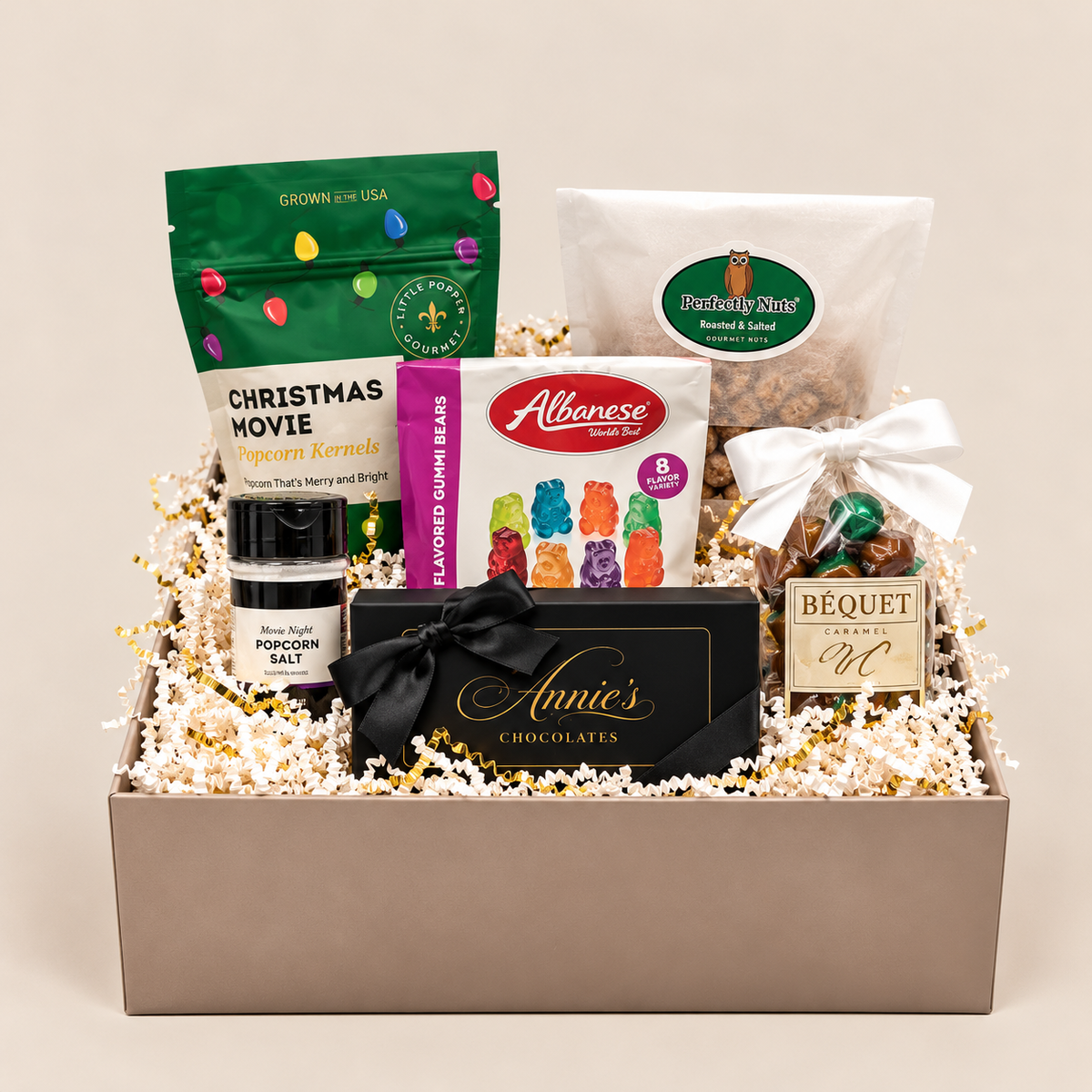 A curated gift basket filled with assorted snacks and treats, including a green bag of Christmas movie popcorn kernels, a white bag of gourmet popcorn, a box of Alton Brown gummy bears, a black box of Annie's Chocolates, a bottle of Blue Moon popcorn salt, and a bag of macaroon bites, arranged in a beige box with shredded paper filler.