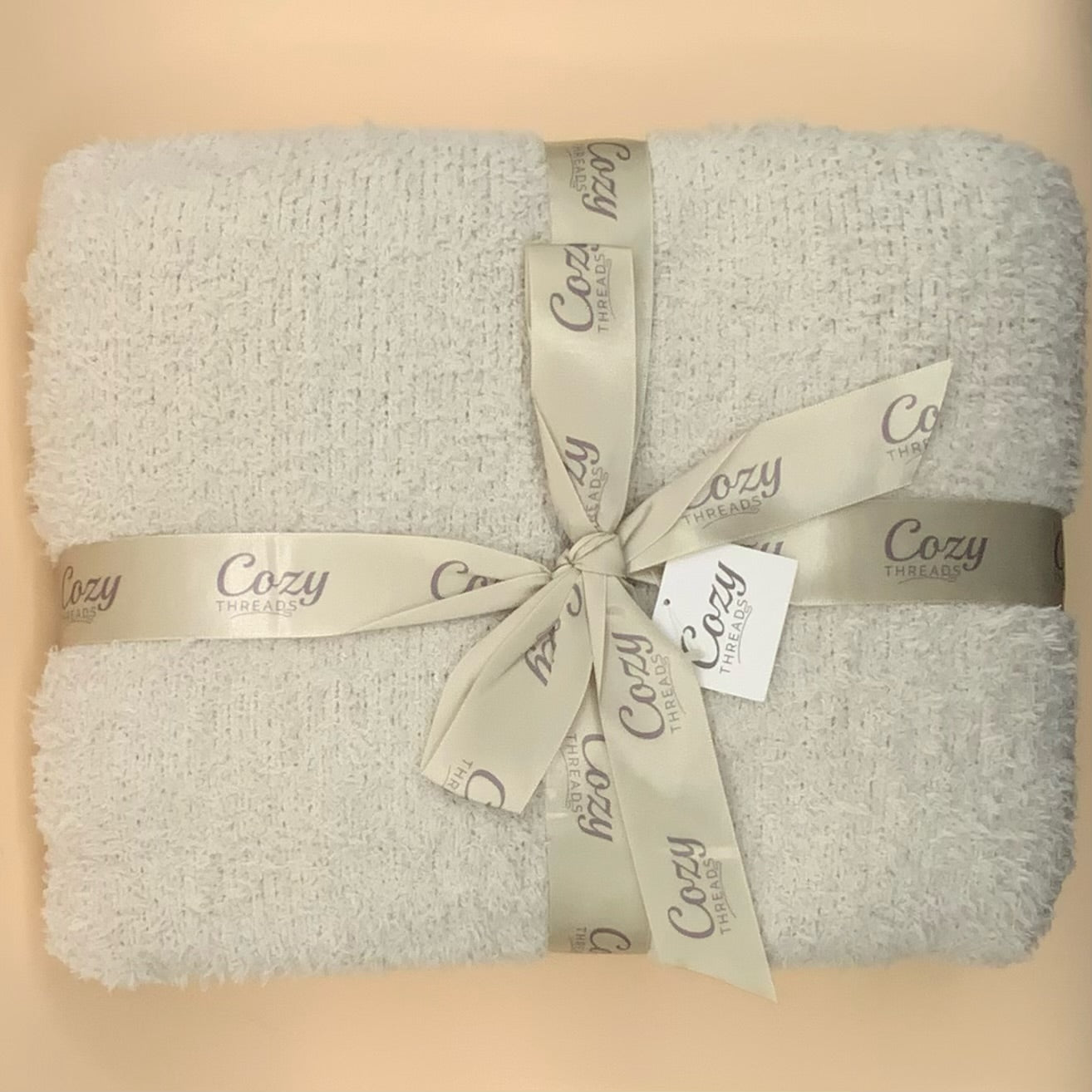 A folded light gray plush bath towel wrapped with a beige Cozy Triplesby ribbon and a small white brand tag, photographed on a warm beige background.