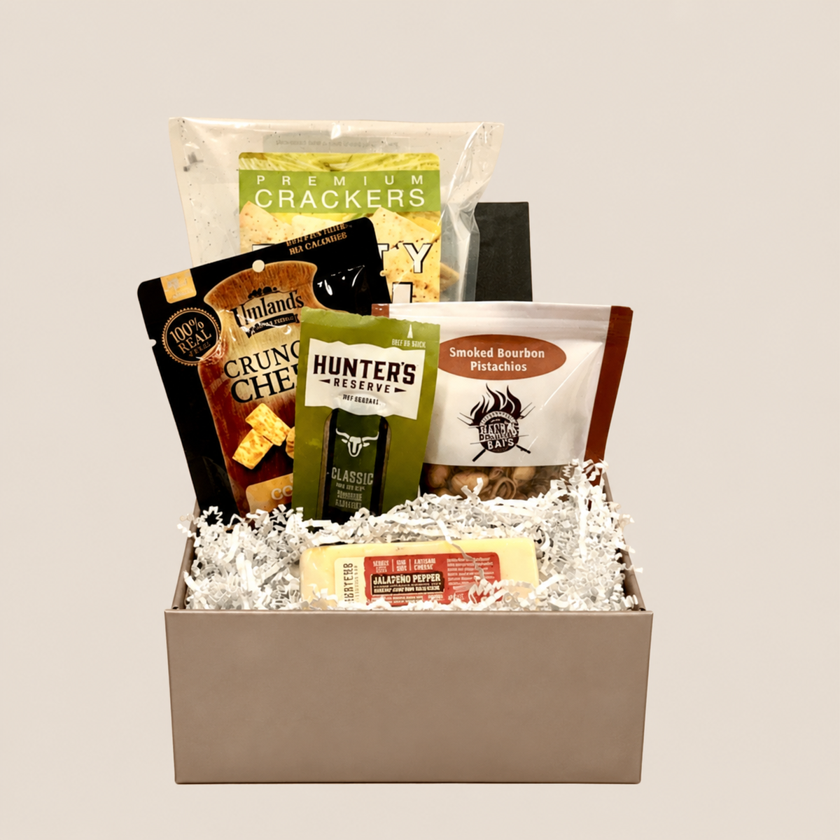 A gourmet snack and cheese gift basket arranged in a metallic silver box with white crinkle paper filler. Visible items include a bag of premium crackers, a package of thinlands Crunch Cheddar, a green bag of Hunters Ranch Classic Cheese, a bag of Smoked Bourbon Pistachios, and a stick of Butterfinger chocolate bar.