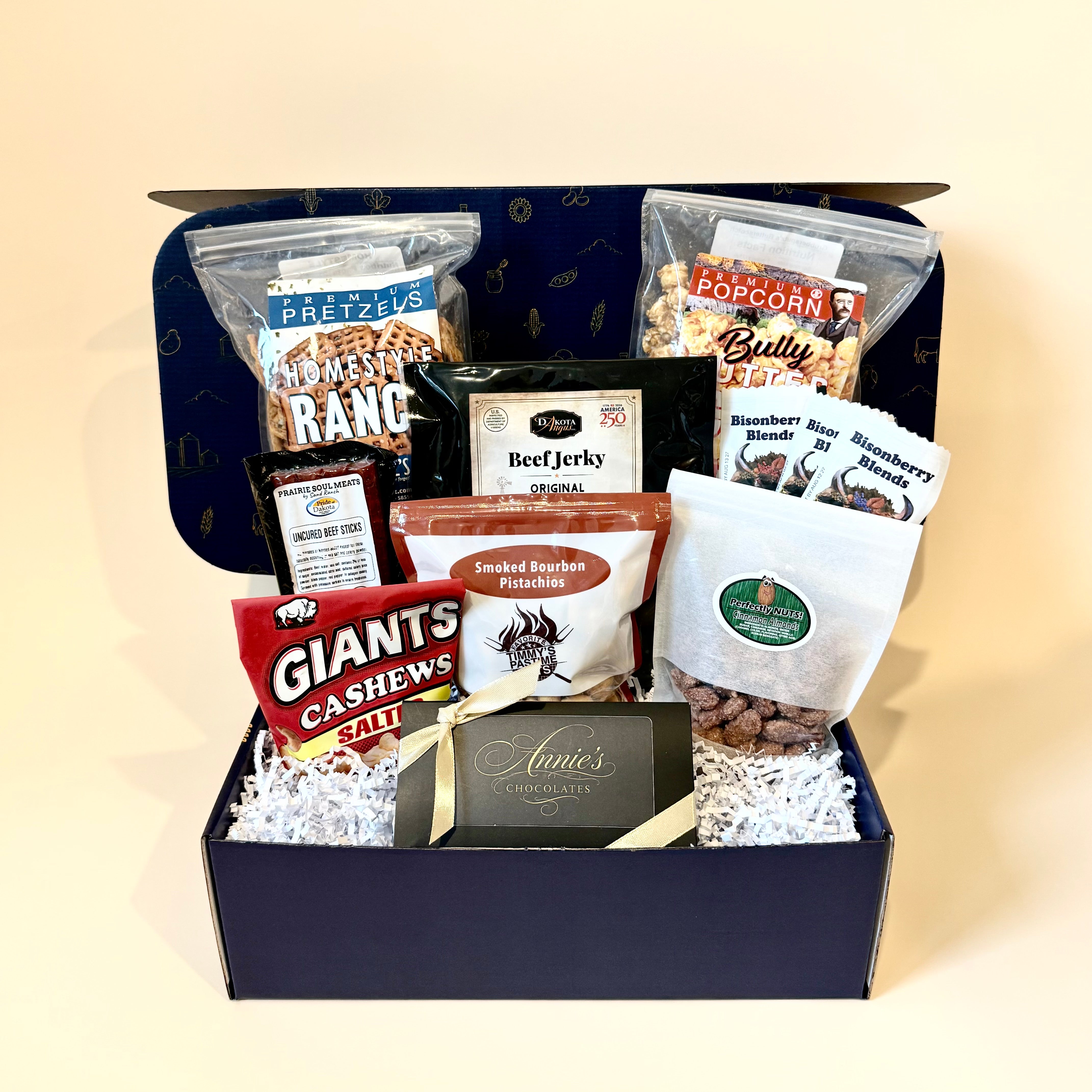 A gourmet snack and coffee gift box arranged in a navy blue box filled with white shredded paper. The assortment includes Pretzels Ranch, Beef Jerky Original, Popcorn Blurry, Smoked Bourbon Pretzels, Raspberry Spice Cinnamon, Giants Cashews Salt, and a card from Annie's Coffee & Tea.