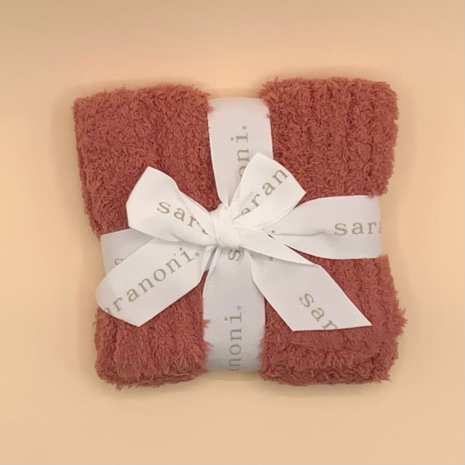 A folded terracotta-colored plush bath towel wrapped with a white ribbon printed with the brand name 'sarani.', set against a solid beige background.