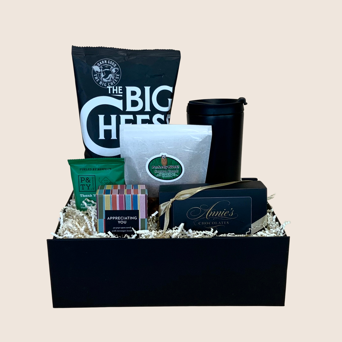 A curated gift basket in a black box filled with assorted packaged snacks and coffee items, including a large bag of The Big Cheese, a green bag of P&Firy chips, a white bag of coffee, a black travel mug, and a small Annie's chocolate box, arranged on shredded paper against a plain beige background.