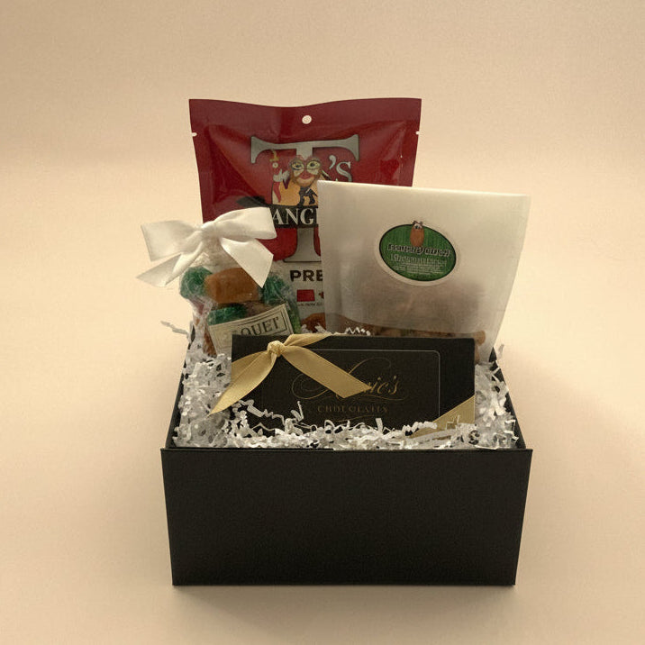 A gift box set containing a black box filled with white crinkle paper, a red pouch, a white bag with a green circular sticker, a small wrapped item tied with a white bow, and a black box tied with a gold ribbon.