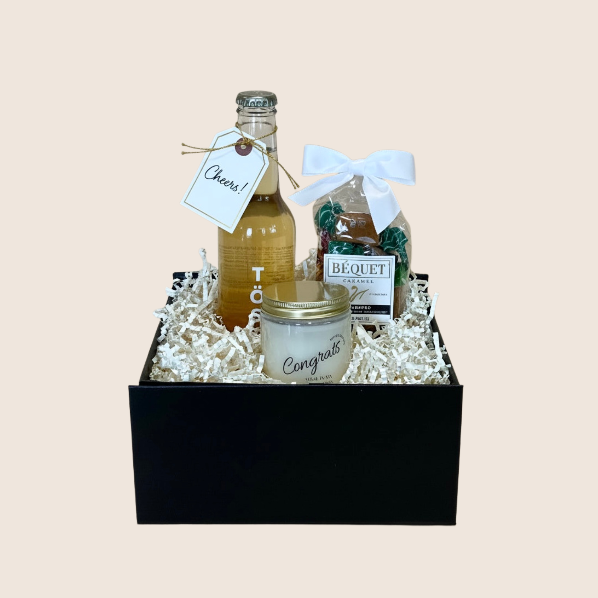 A black gift box filled with white crinkle paper and assorted gift items, including a small bottle of amber liquid with a 'Cheers!' tag, a jar candle reading 'Congrats,' and a bag of Bequet chocolates tied with a white bow.