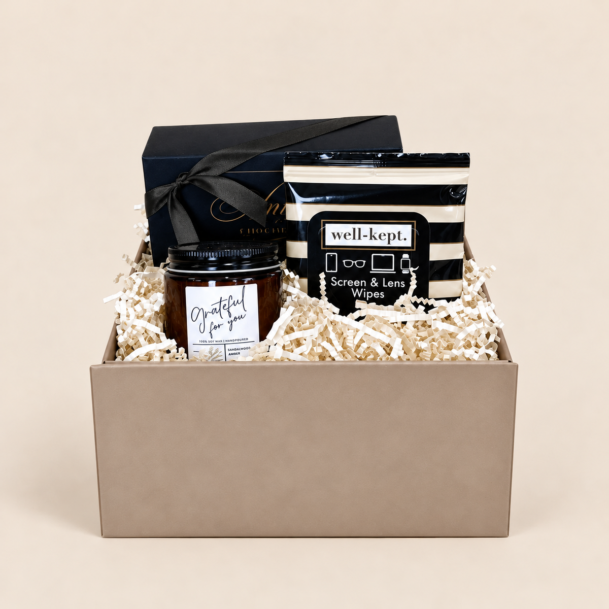 A curated gift box set containing a dark amber candle in a glass jar, a black and white striped screen and lens wipe packet labeled 'well-kept.', and a dark navy gift box tied with a black ribbon, all arranged in beige crinkle paper inside a taupe gift box on a solid beige background.