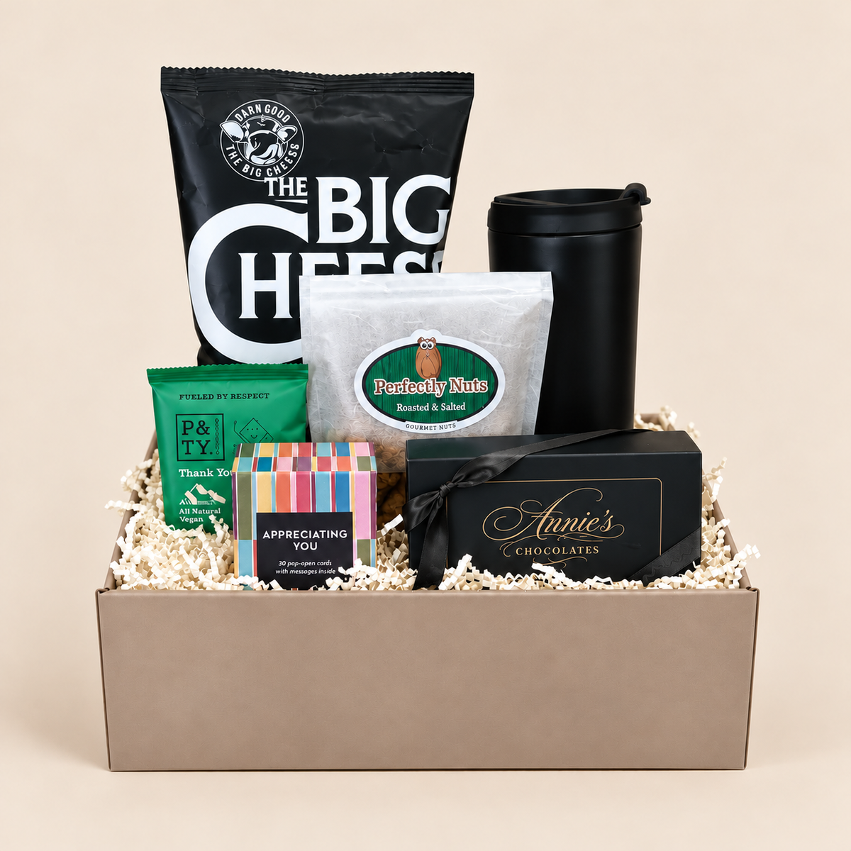 A curated gift basket in a beige box filled with shredded paper, containing a large black bag of The Big Chefs snacks, a black travel mug, a white packet of Rooty Nite, a green packet of Fisher Rye Breakfast, a colorful bar box, and a black box of Annie's Chocolates.