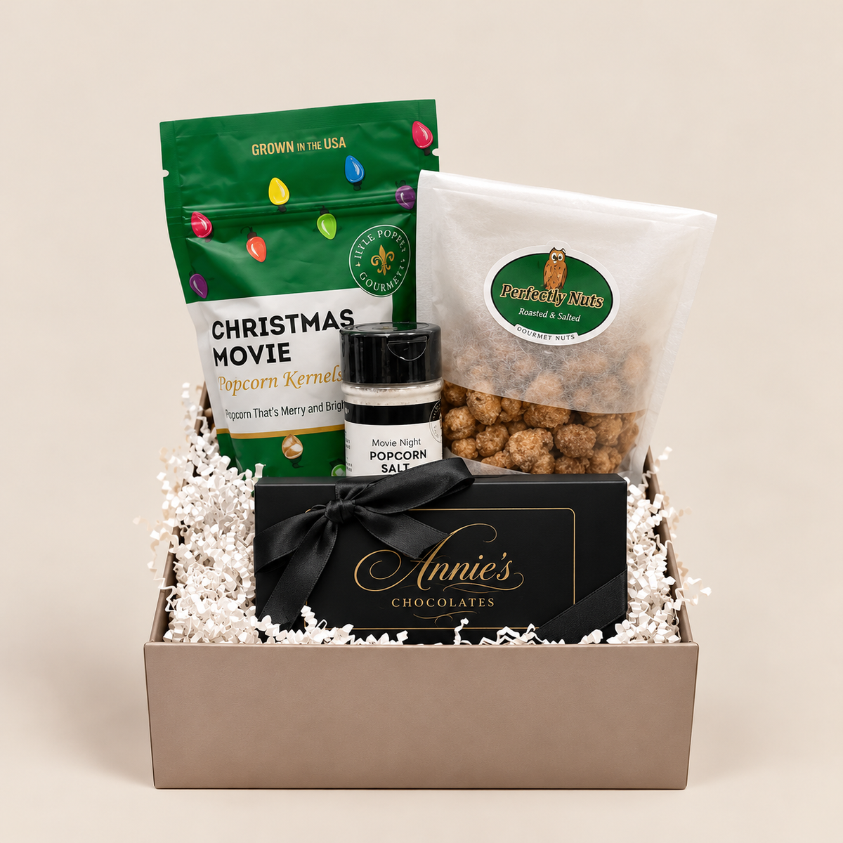 A gift basket filled with assorted snacks and chocolates, including a green bag of Christmas Movie Popcorn Kernel, a clear bag of Peanut Butter Chips, a jar of Minnie Night Popcorn Salt, and a black box of Annie's Chocolates tied with a black ribbon, arranged in white paper shred inside a beige basket on a light beige background.