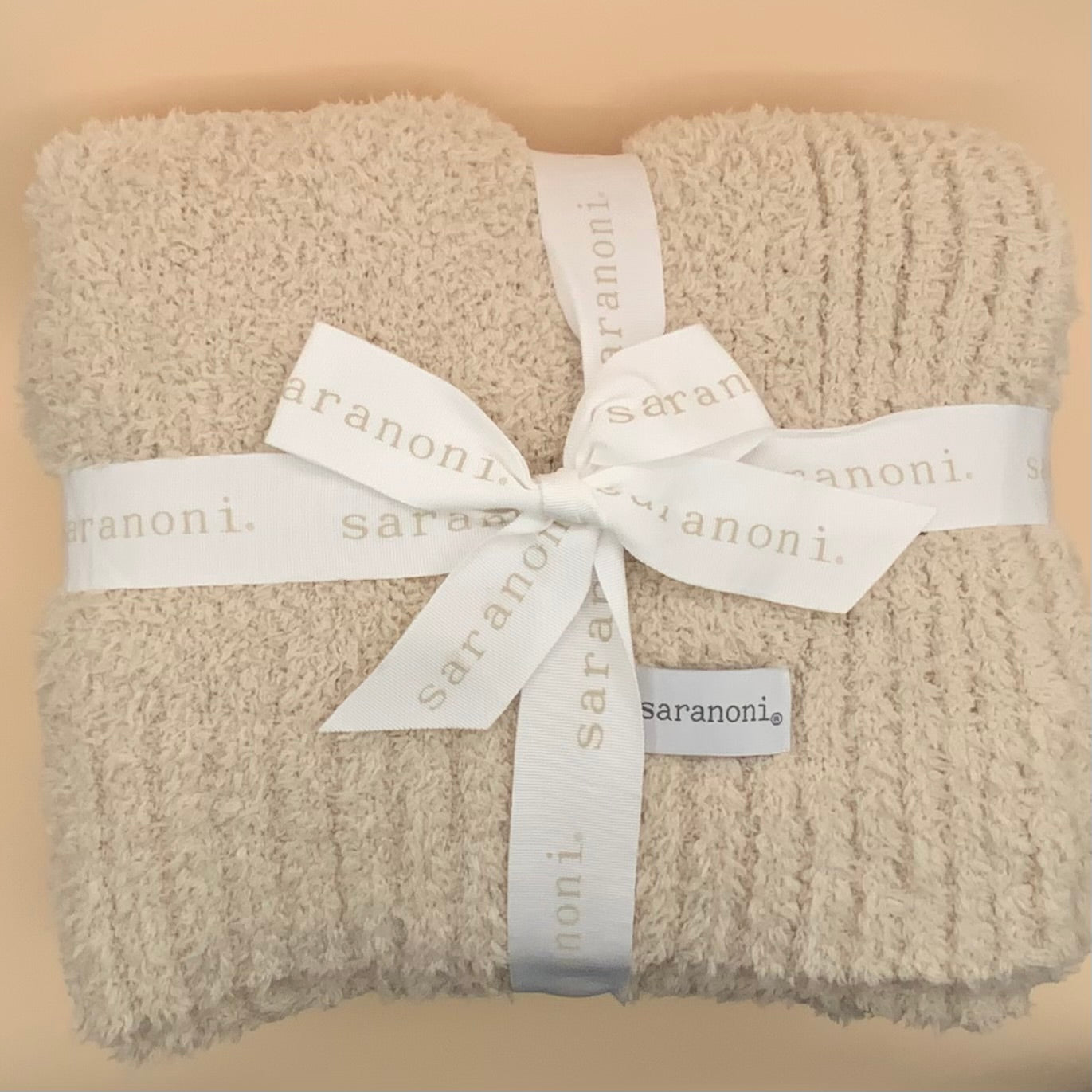 A folded cream-colored textured knit blanket or throw, wrapped with a white satin ribbon printed with the brand name 'saranoni'.