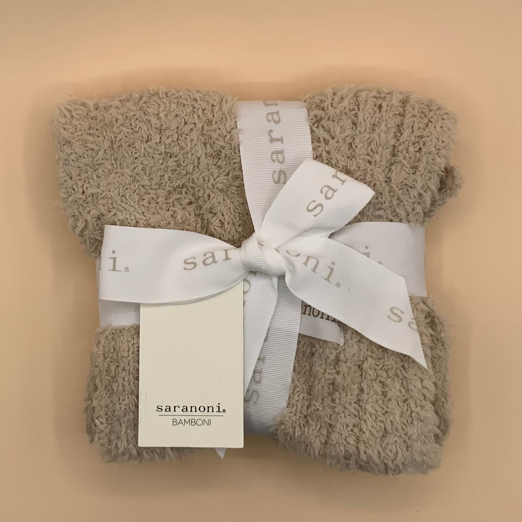 A folded beige bamboo towel or blanket wrapped with a white satin ribbon printed with the brand name 'saranoni.' and a matching paper tag, set against a solid beige background.