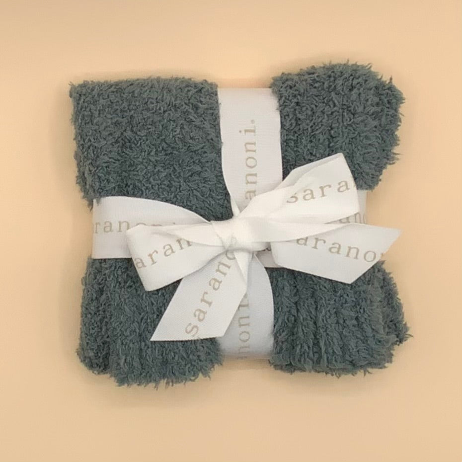 A folded gray terry cloth towel wrapped with a white satin ribbon printed with the brand name 'sarah anon.' in a light gray font, set against a solid pale beige background.