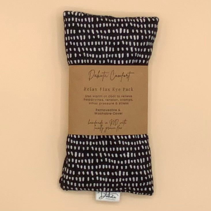 A packaged Debito Comfort Relax Flax Eye Pack, showing the black and white patterned eye pillow with a brown paper band label in the center, set against a solid beige background.